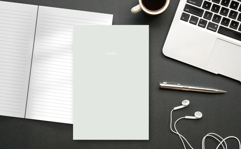 sea sand gray notebook cover with the word 'cunt.', inside pages on a desk with a lap top, coffee, pens and ear buds.