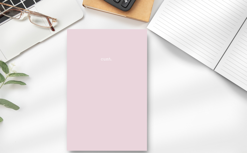 Soft pink notebook cover with subtle image of the word cunt. on a desk with a laptop, calculator, glasses, plant and the inside lined pages.