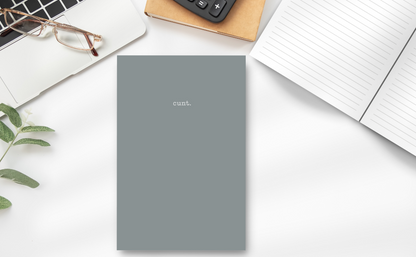 This image features a sleek, granite-grey notebook with the word "cunt." subtly printed at the top. On a desk showing the inside lined pages, laptop, calculator and plant.