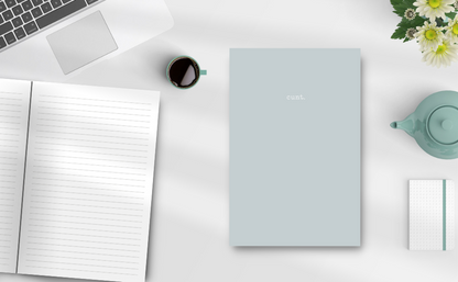 Sage grey colour notebook with the word cunt. on a desk showing the inside pages, keyboard, tea pot, flowers