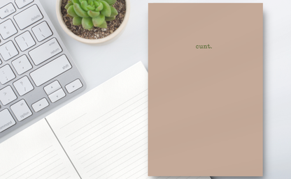 Notebook with 'cunt.' on a desk next to a keyboard and plant