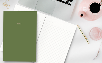 Green notebook with 'cunt' on a desk with a laptop and a tea pot.