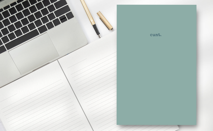Notebook with 'cunt.' on a desk next to a laptop